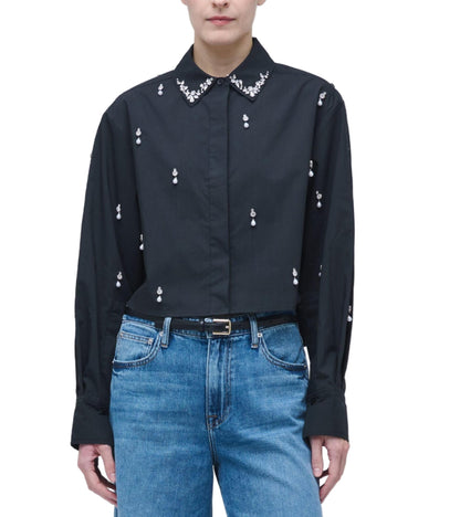 A person wearing the Simkhai Renata Embellished Cropped Shirt with small embroidered details and a beaded collar, paired with blue jeans and a black belt.