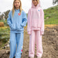 Two young girls stand outdoors on a dirt path, one wearing a blue relaxed fit Feather 4 Arrow Girls’ Cozy Sweatpant set and the other in a pink hoodie and Feather 4 Arrow Girls’ Cozy Sweatpant set. Both have long blonde hair and neutral expressions.