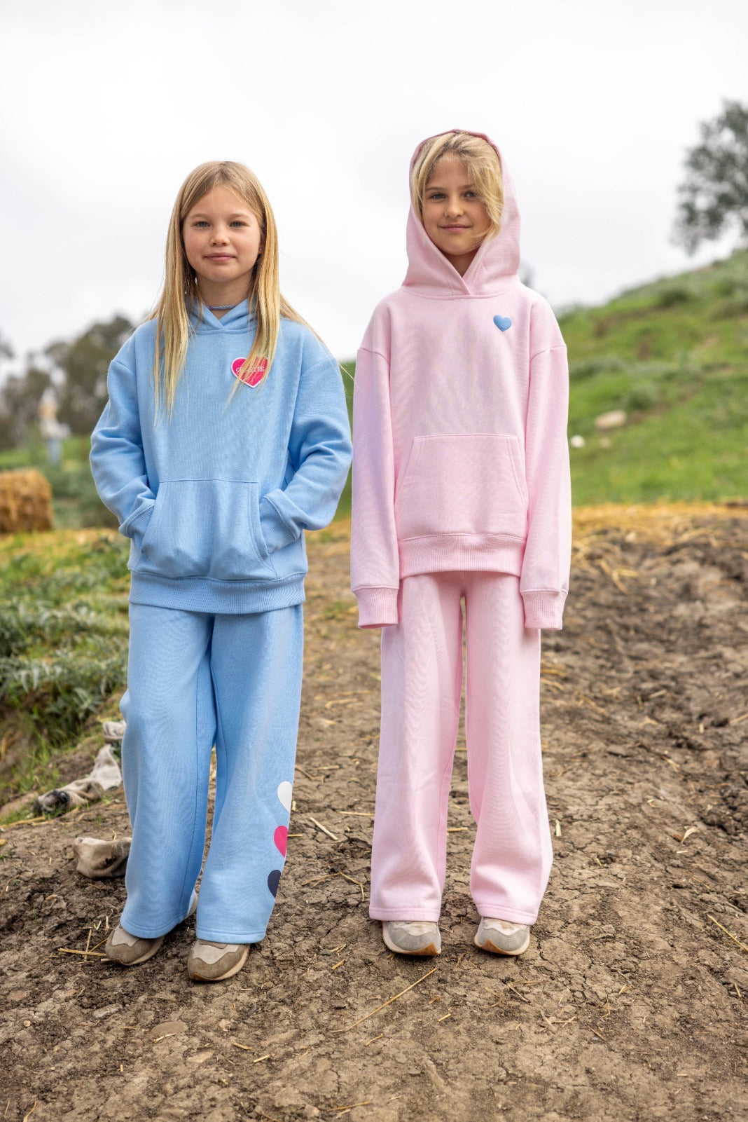 Two young girls stand outdoors on a dirt path, one wearing a blue relaxed fit Feather 4 Arrow Girls’ Cozy Sweatpant set and the other in a pink hoodie and Feather 4 Arrow Girls’ Cozy Sweatpant set. Both have long blonde hair and neutral expressions.