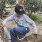Wearing the Feather 4 Arrow Boys’ Wyatt Jogger with an adjustable waistband, a child in everyday wear—including a cap and long sleeves—climbs a tree and looks down, surrounded by greenery and dirt. The jogger provides comfort and flexibility for every outdoor adventure.