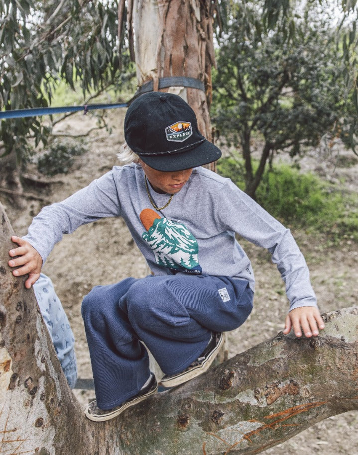 Wearing the Feather 4 Arrow Boys’ Wyatt Jogger with an adjustable waistband, a child in everyday wear—including a cap and long sleeves—climbs a tree and looks down, surrounded by greenery and dirt. The jogger provides comfort and flexibility for every outdoor adventure.