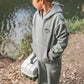 A young boy in a gray hoodie and Feather 4 Arrow Boys’ Wyatt Jogger with an adjustable waistband stands by a riverbank, holding a fishing tackle box and gazing at the water—making these joggers perfect for everyday outdoor adventures.