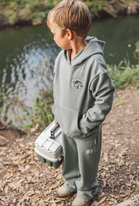A young boy in a gray hoodie and Feather 4 Arrow Boys’ Wyatt Jogger with an adjustable waistband stands by a riverbank, holding a fishing tackle box and gazing at the water—making these joggers perfect for everyday outdoor adventures.