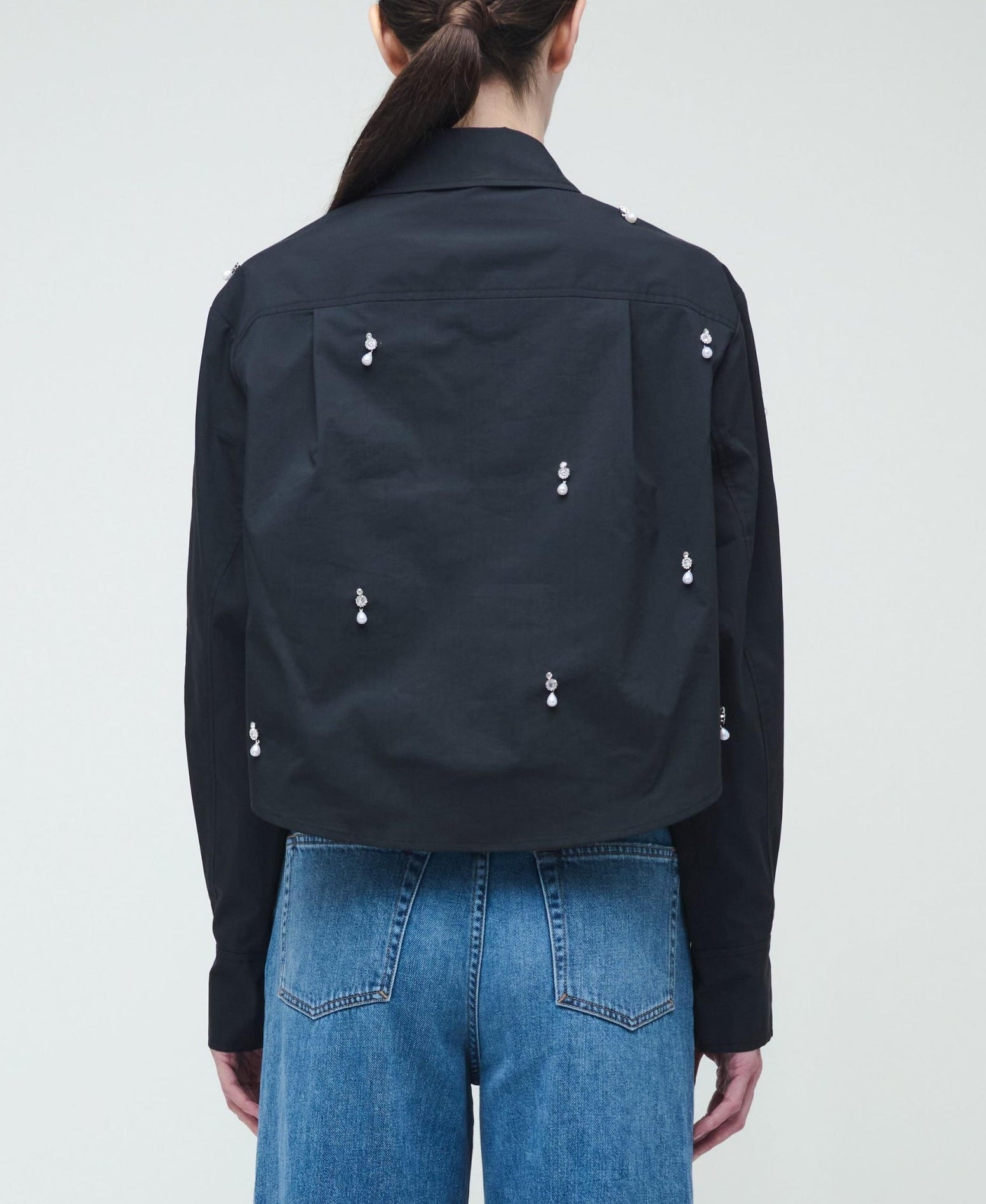 A person is seen from behind wearing the Simkhai Renata Embellished Cropped Shirt, featuring crystal pearl details on the back, paired with blue jeans against a plain background.
