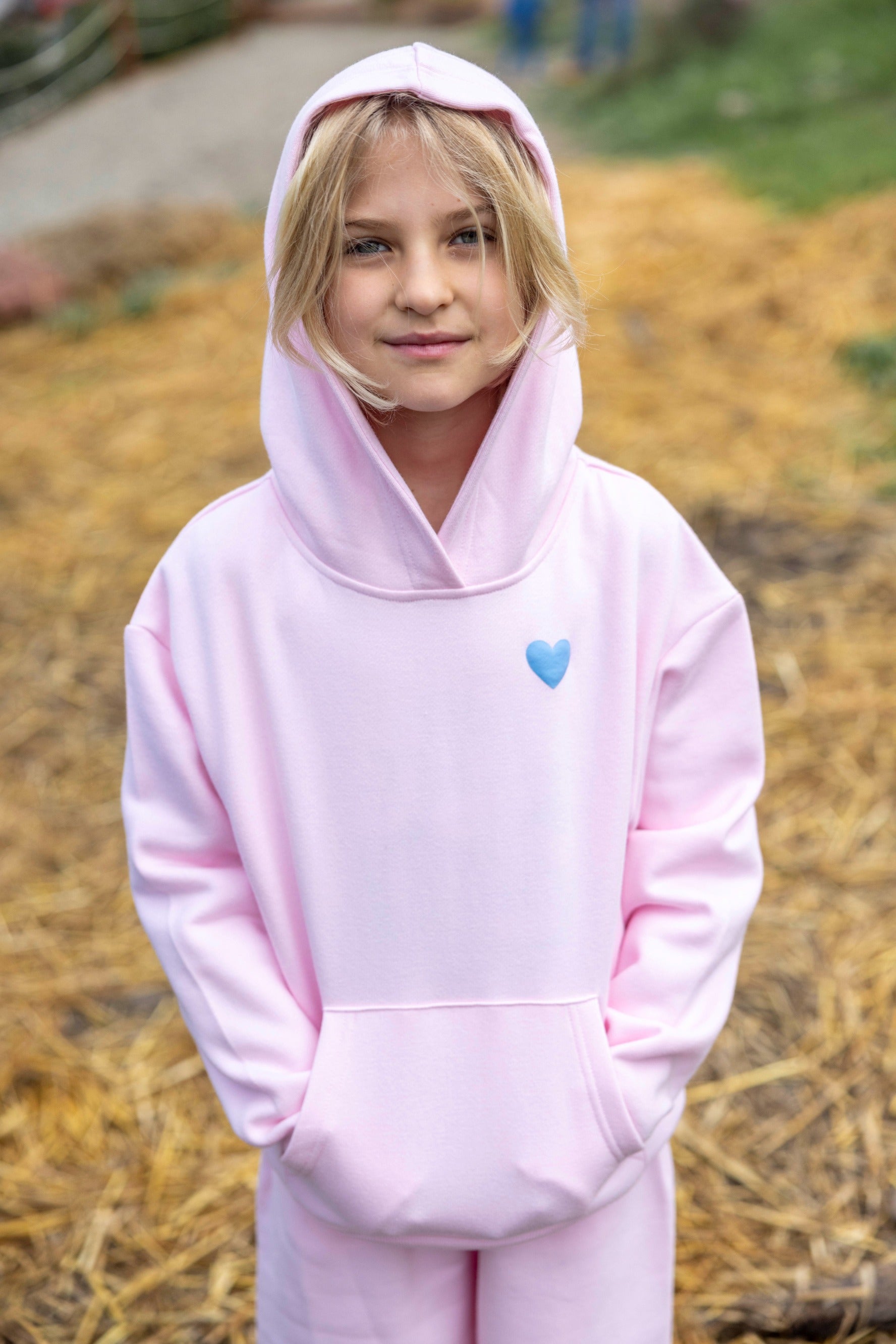 A child stands outdoors wearing the Feather 4 Arrow Girls’ Hadley Cozy Hoodie in light pink, featuring a blue heart design, with hands in pockets and looking at the camera.