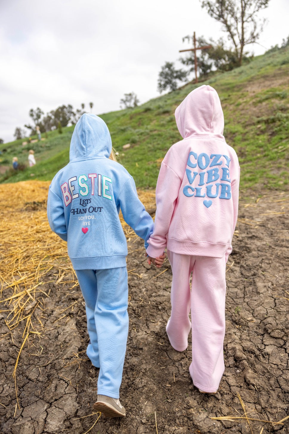 Two children stroll hand in hand outdoors, each bundled up in a plush Feather 4 Arrow hoodie that guarantees comfort on every outing. One wears the Girls’ Hadley Bestie Hoody with “BESTIE” proudly displayed, while the other sports a “COZY VIBE CLUB” design. The path beneath them is cracked and scattered with straw.