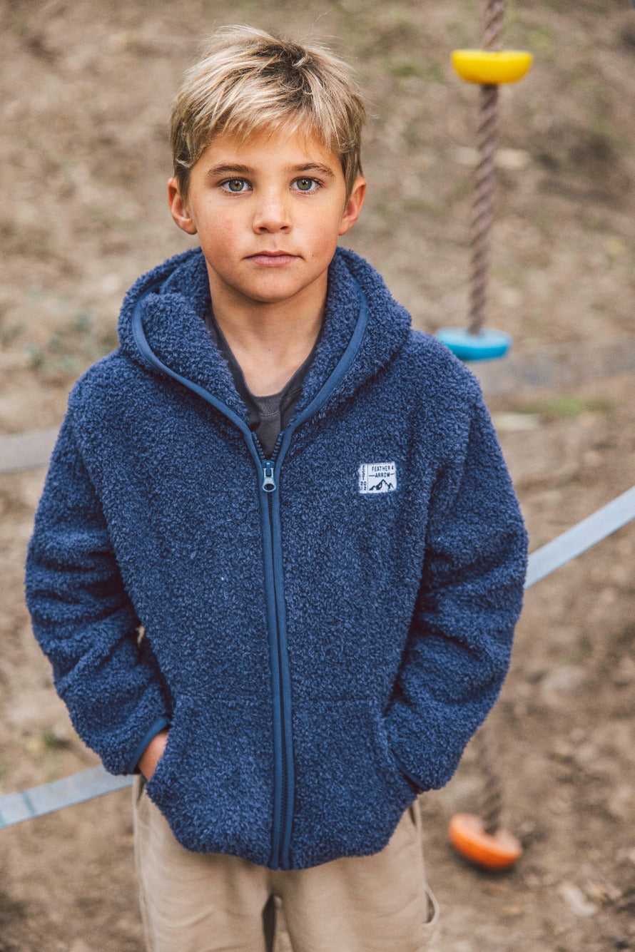 A young boy wears the Feather 4 Arrow Baby Boys’ and Boys’ Rocky Sherpa Jacket as he stands outdoors with his hands in his pockets, looking at the camera. Colorful climbing holds and a rope appear in the background, adding a playful touch to this cozy jacket’s casual style.
