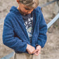 A young child wearing the Feather 4 Arrow Baby Boys’ and Boys’ Rocky Sherpa Jacket in blue with beige pants is looking down and zipping up their jacket outdoors.
