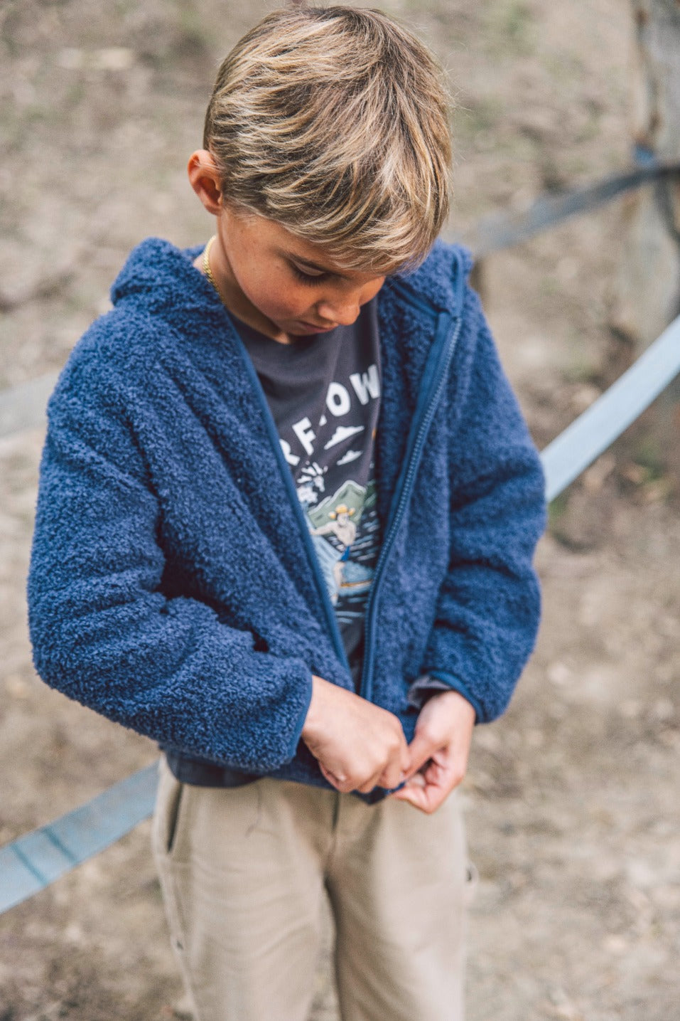 A young child wearing the Feather 4 Arrow Baby Boys’ and Boys’ Rocky Sherpa Jacket in blue with beige pants is looking down and zipping up their jacket outdoors.
