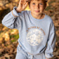 A young child wearing the Feather 4 Arrow Baby Boys’ and Boys’ Ryder Outside Sweatshirt in light blue and a tan cap stands outdoors on fallen leaves, holding the brim of his hat. The sweatshirt features the phrase “I’D RATHER BE OUTSIDE,” combining comfort with timeless style.