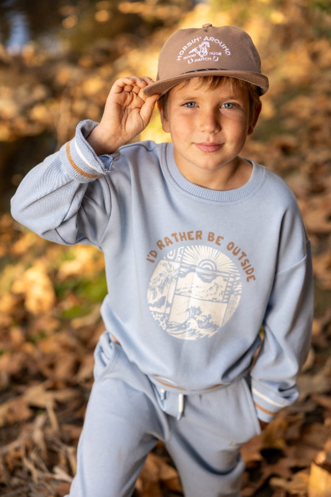 A young child wearing the Feather 4 Arrow Baby Boys’ and Boys’ Ryder Outside Sweatshirt in light blue and a tan cap stands outdoors on fallen leaves, holding the brim of his hat. The sweatshirt features the phrase “I’D RATHER BE OUTSIDE,” combining comfort with timeless style.