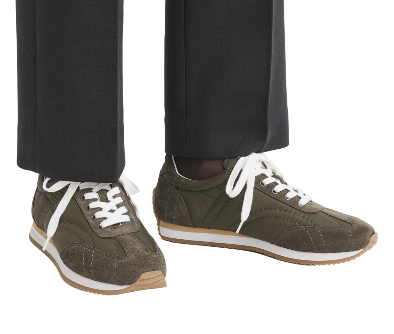 A person wearing black trousers and Toteme Sport Sneakers in olive green with white laces, gum soles, and subtle suede trims.