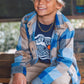A smiling boy wears the Feather 4 Arrow Boys’ Surf Club Long Sleeved Tee, paired with a blue plaid jacket, navy cap, and khaki pants as he sits on a wooden bench in front of a blurred chalkboard background—a perfect look for kids’ everyday wear.