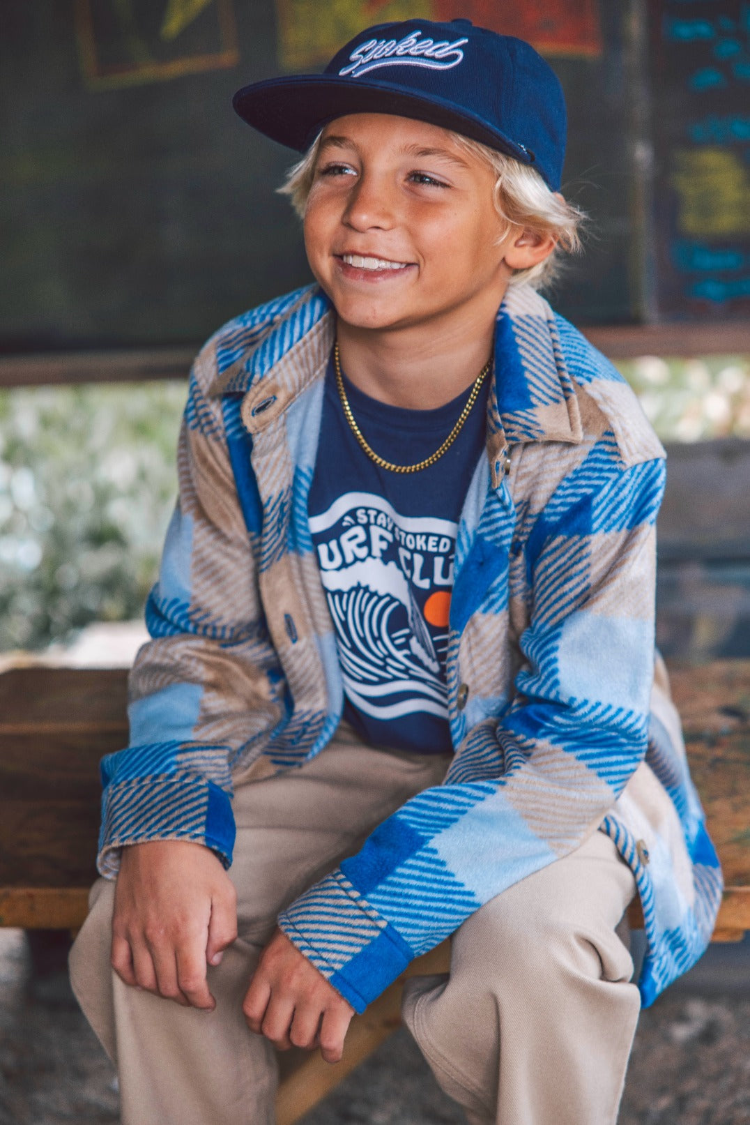 A smiling boy wears the Feather 4 Arrow Boys’ Surf Club Long Sleeved Tee, paired with a blue plaid jacket, navy cap, and khaki pants as he sits on a wooden bench in front of a blurred chalkboard background—a perfect look for kids’ everyday wear.