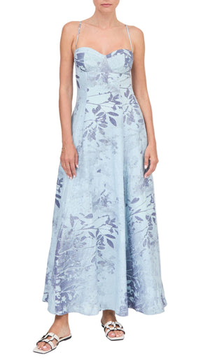 A woman wearing the Simkhai Rudie Dress, a light blue, ankle-length, sleeveless dress with a subtle leaf pattern and thin straps, paired with white sandals.