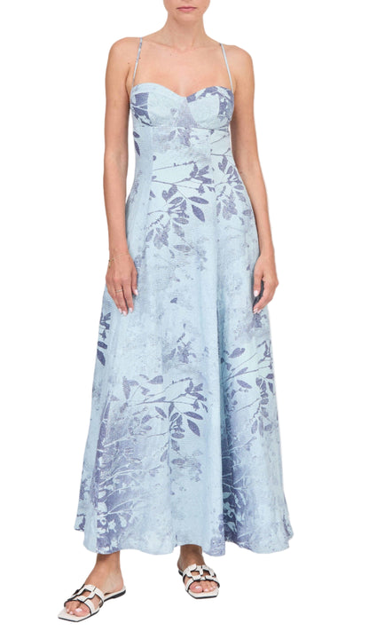 A woman wearing the Simkhai Rudie Dress, a light blue, ankle-length, sleeveless dress with a subtle leaf pattern and thin straps, paired with white sandals.