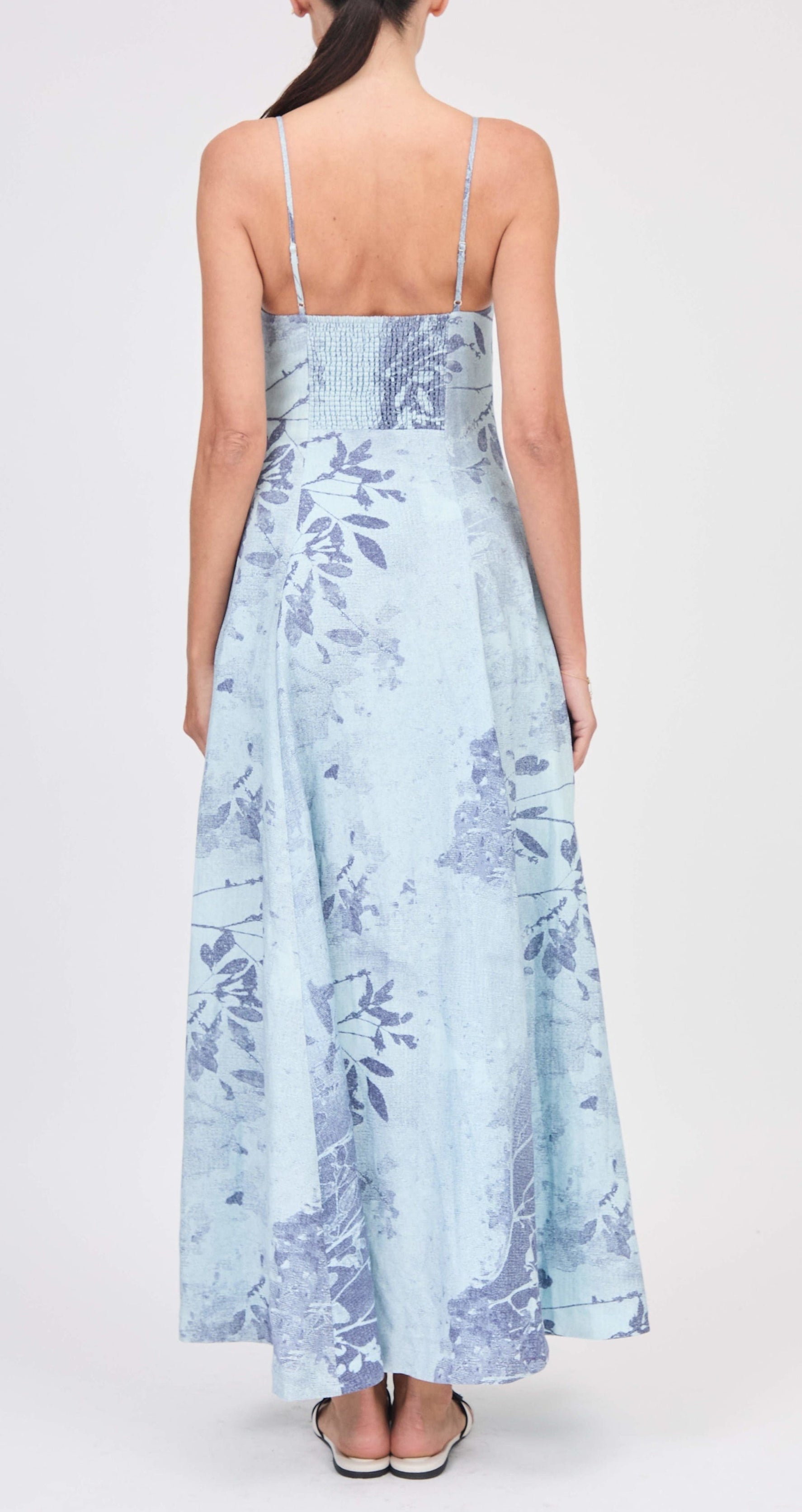 A woman is shown from the back wearing the Simkhai Rudie Dress—a light blue, sleeveless, ankle-length floral dress—against a plain background.