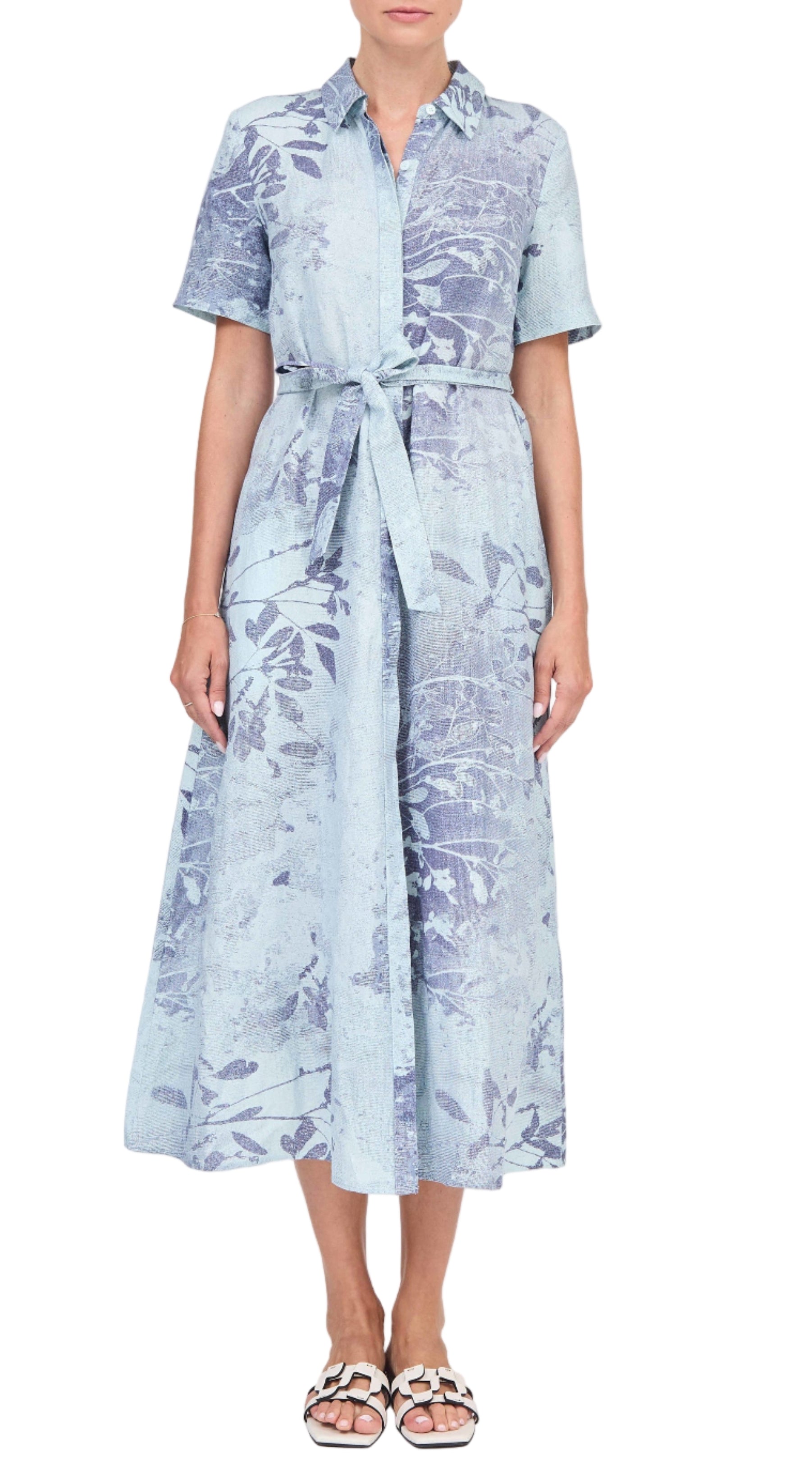 A woman wears the Simkhai Audria Belted Dress, a light blue, short-sleeved, collared midi dress with a tied waist and floral pattern, styled with white flat sandals.