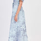 A woman stands in profile wearing the Simkhai Audria Belted Dress—a light blue, short-sleeved midi dress with a subtle floral pattern—paired with white sandals.