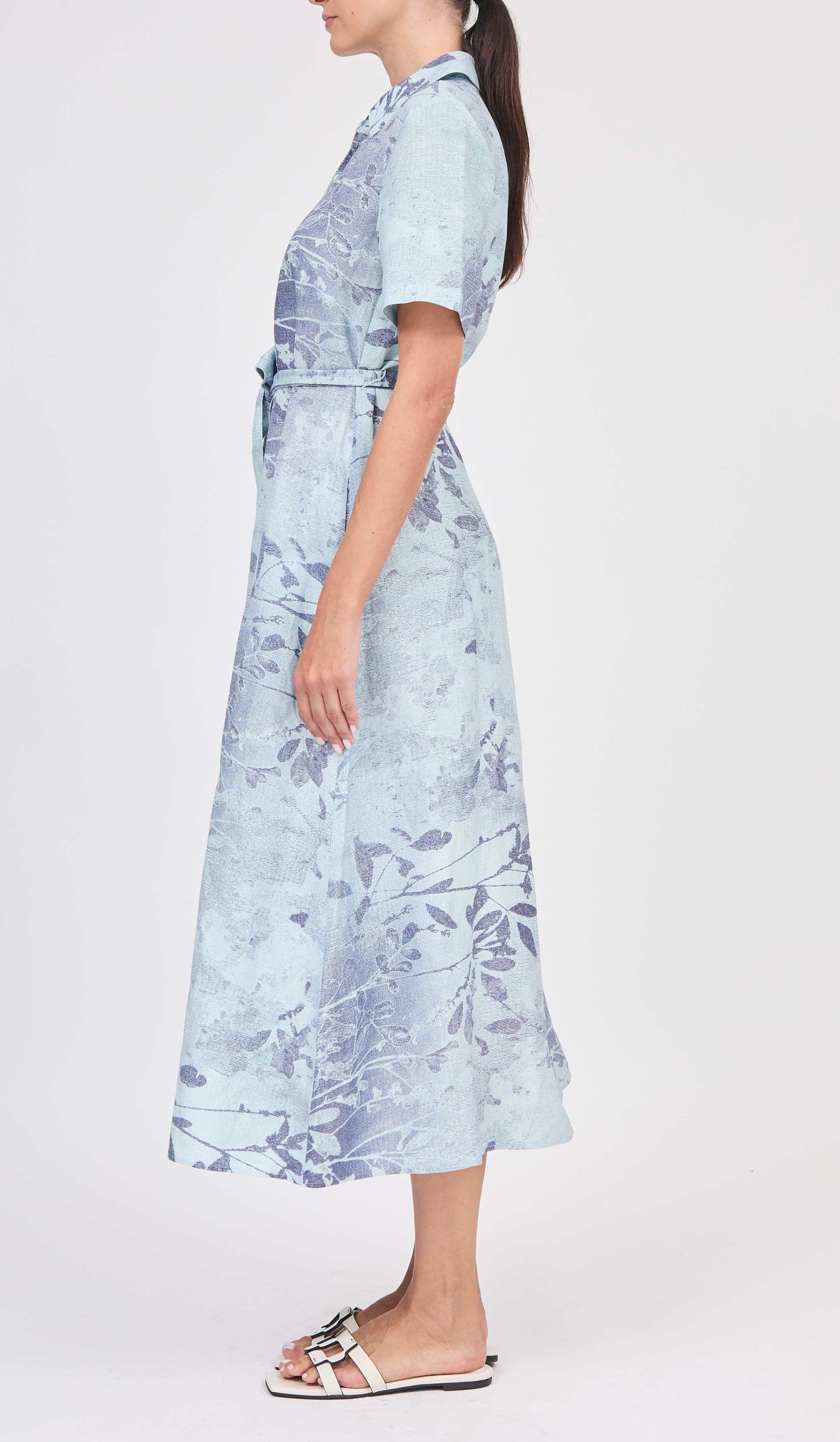 A woman stands in profile wearing the Simkhai Audria Belted Dress—a light blue, short-sleeved midi dress with a subtle floral pattern—paired with white sandals.