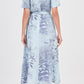 A woman stands with her back to the camera, wearing the Simkhai Audria Belted Dress—a light blue, short-sleeve, collared, midi dress with a botanical print—paired with flat sandals.
