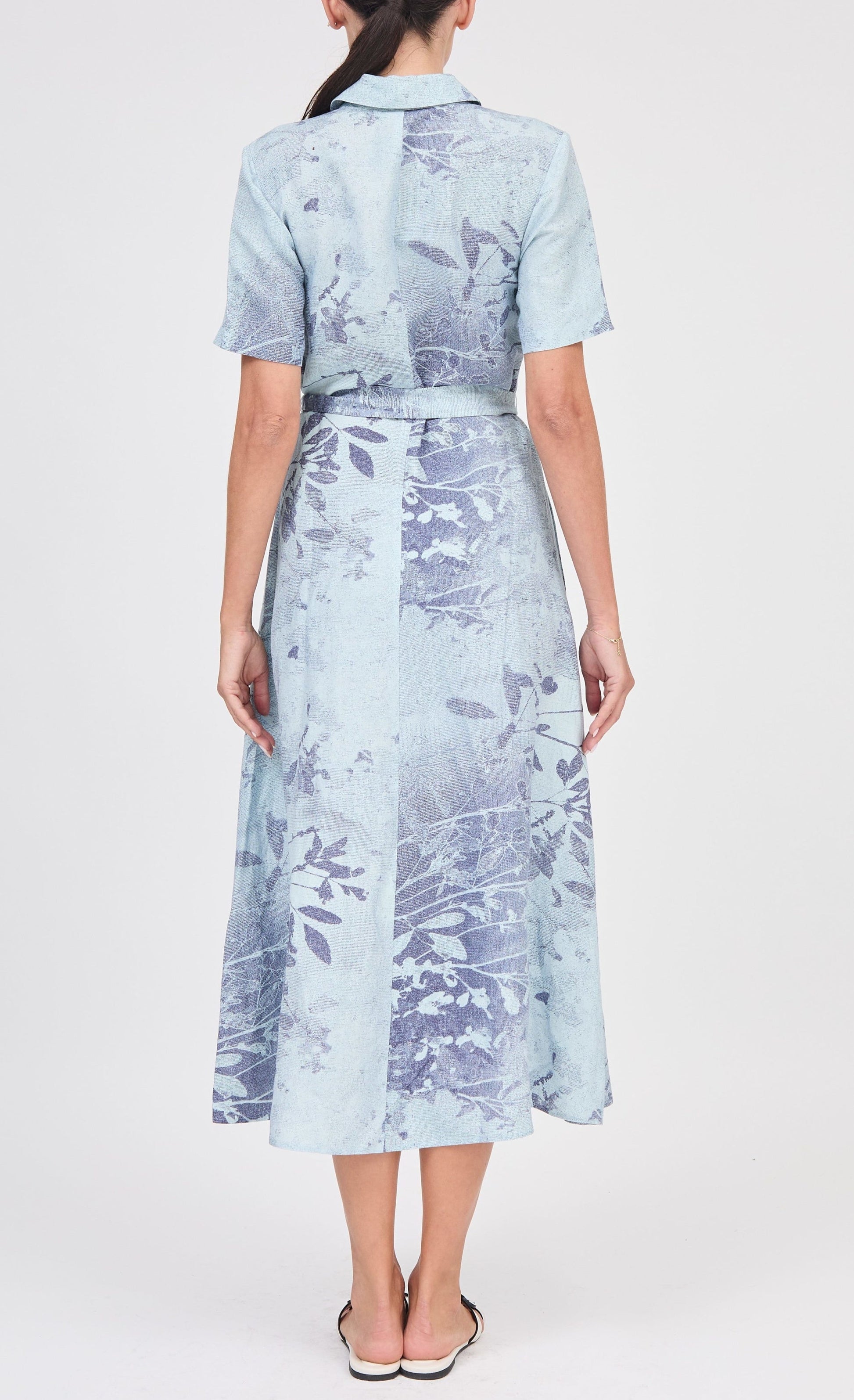 A woman stands with her back to the camera, wearing the Simkhai Audria Belted Dress—a light blue, short-sleeve, collared, midi dress with a botanical print—paired with flat sandals.