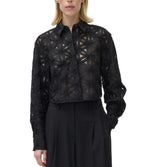 A woman wears the Simkhai Anders Shirt, a black sheer blouse with a geometric floral pattern and long sleeves, paired with high-waisted black trousers.