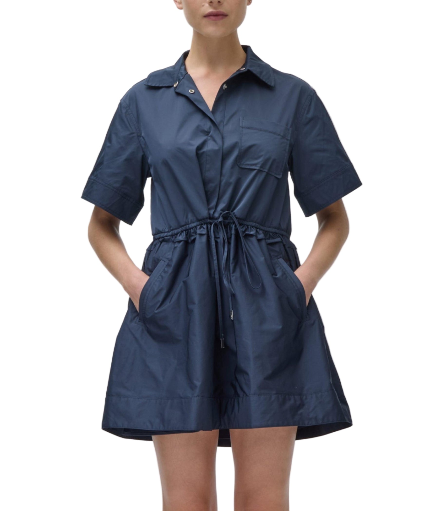 A person wearing the Simkhai Dawson Shirt Dress, a navy blue short-sleeve button-up style with a drawstring waist and side pockets, stands against a plain background.