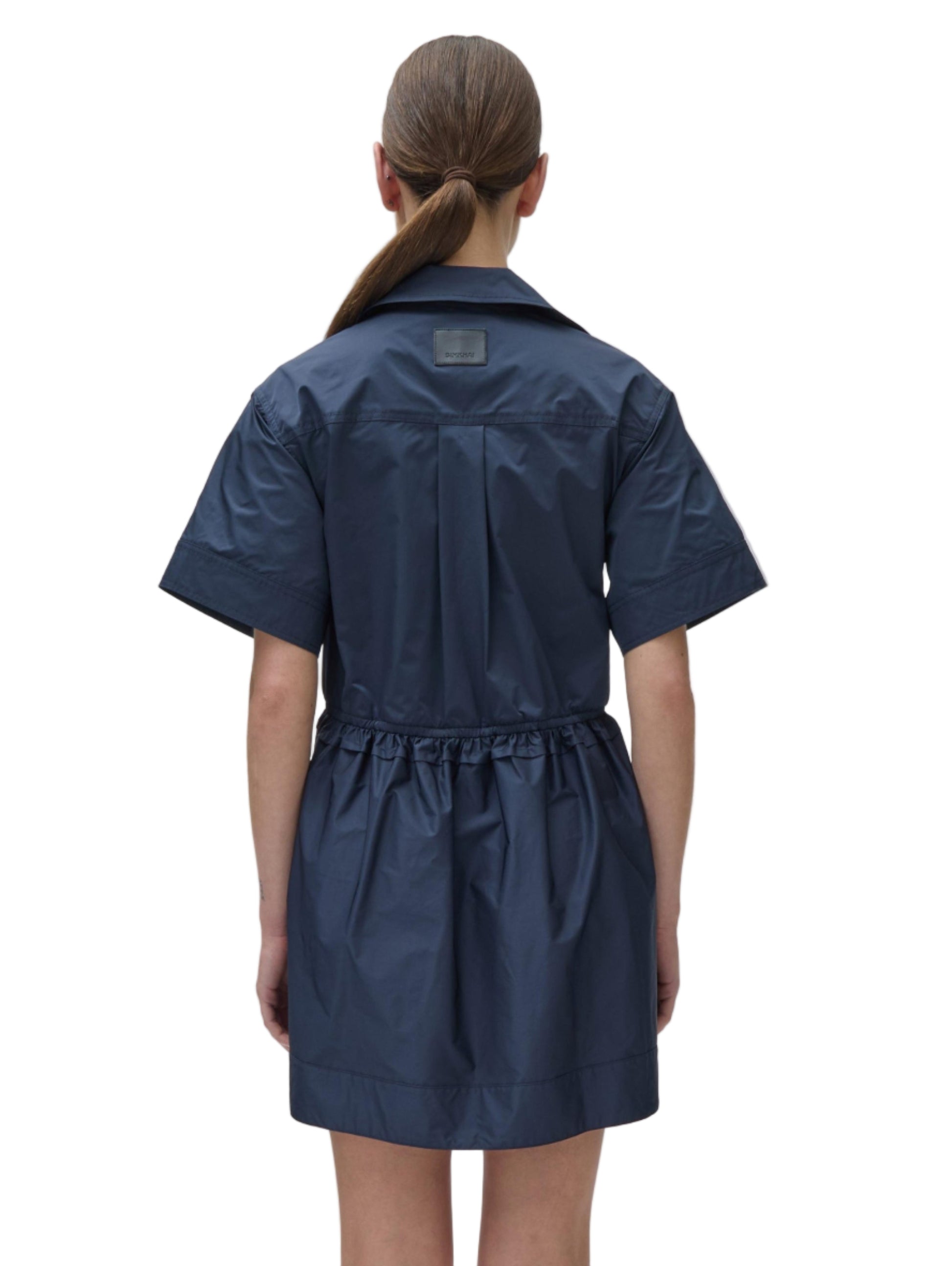Person facing away, wearing the Simkhai Dawson Shirt Dress in navy blue. The dress features short sleeves, a collared neckline, and an elastic waist. Hair is tied back in a low ponytail.