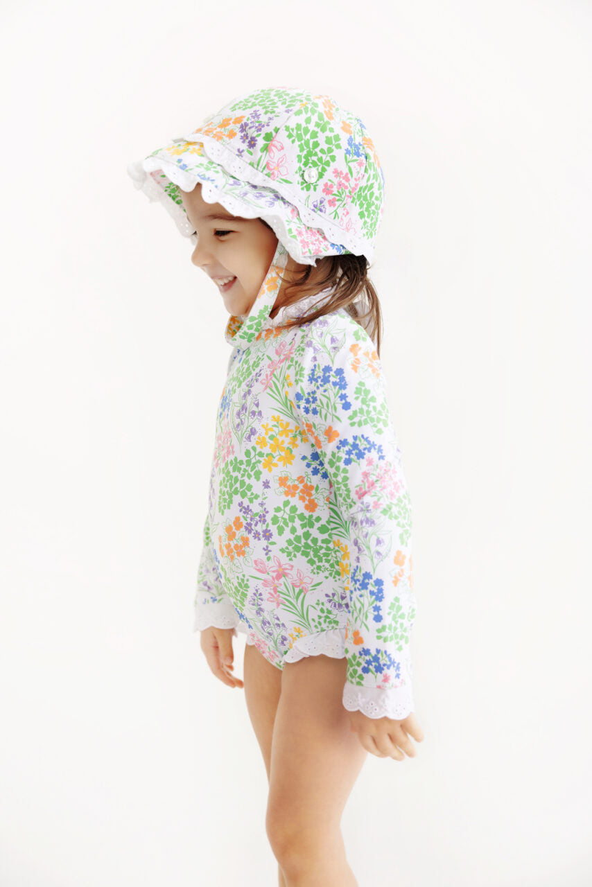 A young child stands smiling against a plain white background, wearing The Beaufort Bonnet Company’s Beaufort Swim Bucket Hat—a sun-ready accessory—paired with a matching long-sleeve, swim-ready nylon spandex swimsuit in a floral pattern.
