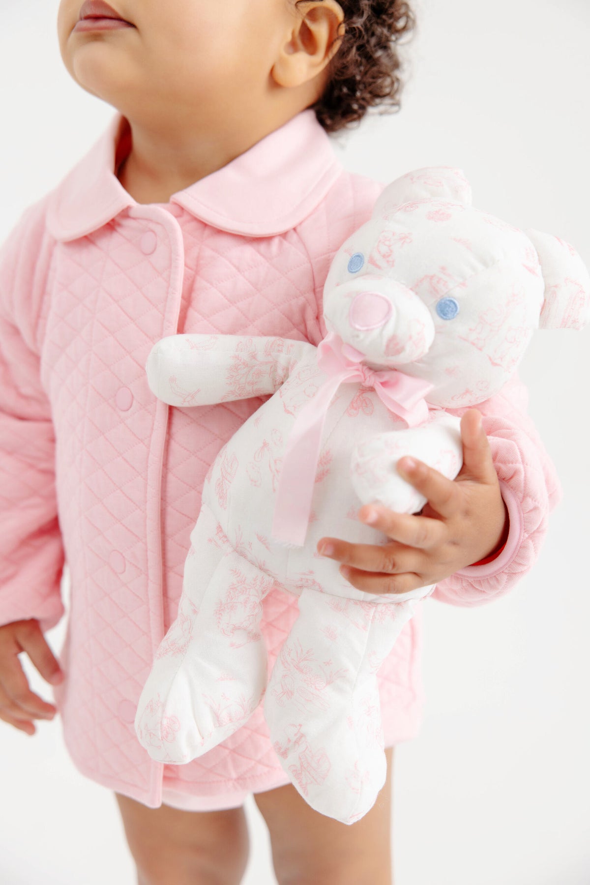 A young child in a pink quilted jacket holds the Bradley Bear - Sir Propers Sweet Arrival, a white teddy bear with pink patterns and a pink ribbon from The Beaufort Bonnet Company—an adorable companion that would make the perfect baby shower gift.