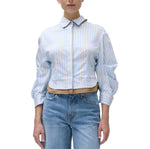 A person wearing the Simkhai Boone Combo Shirt, a light blue and white striped long-sleeve blouse with puffed sleeves, paired with blue jeans and standing against a white background.