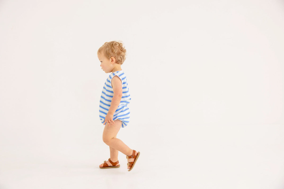 A toddler wearing The Beaufort Bonnet Company’s Baby Boys’ Broadcloth Benjamin Bubble—a blue and white striped romper—and brown sandals walks to the left against a plain white background.