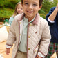 A young boy smiles at the camera outdoors, wearing The Beaufort Bonnet Company’s Baby Boys’ and Boys’ Caldwell Quilted Coat in beige with a corduroy collar, paired with Keeneland Khaki pants; other children and greenery are visible in the background.