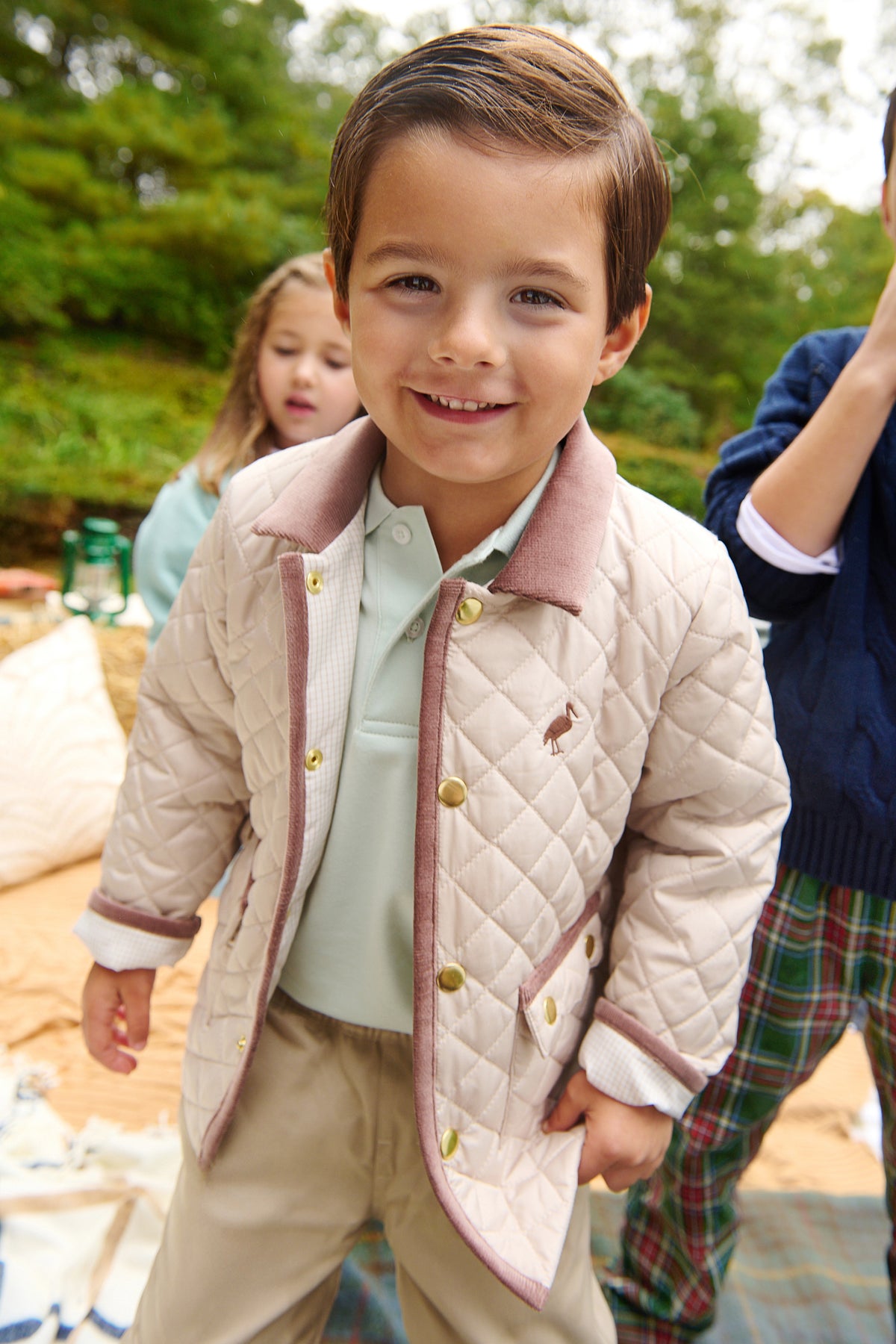 A young boy smiles at the camera outdoors, wearing The Beaufort Bonnet Company’s Baby Boys’ and Boys’ Caldwell Quilted Coat in beige with a corduroy collar, paired with Keeneland Khaki pants; other children and greenery are visible in the background.