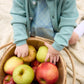 A young child in a blue outfit sits on a blanket, cozily wrapped in The Beaufort Bonnet Company’s Baby Girls’ and Girls’ Cambridge Cardigan with pearlized buttons, while holding a basket filled with red and green apples.
