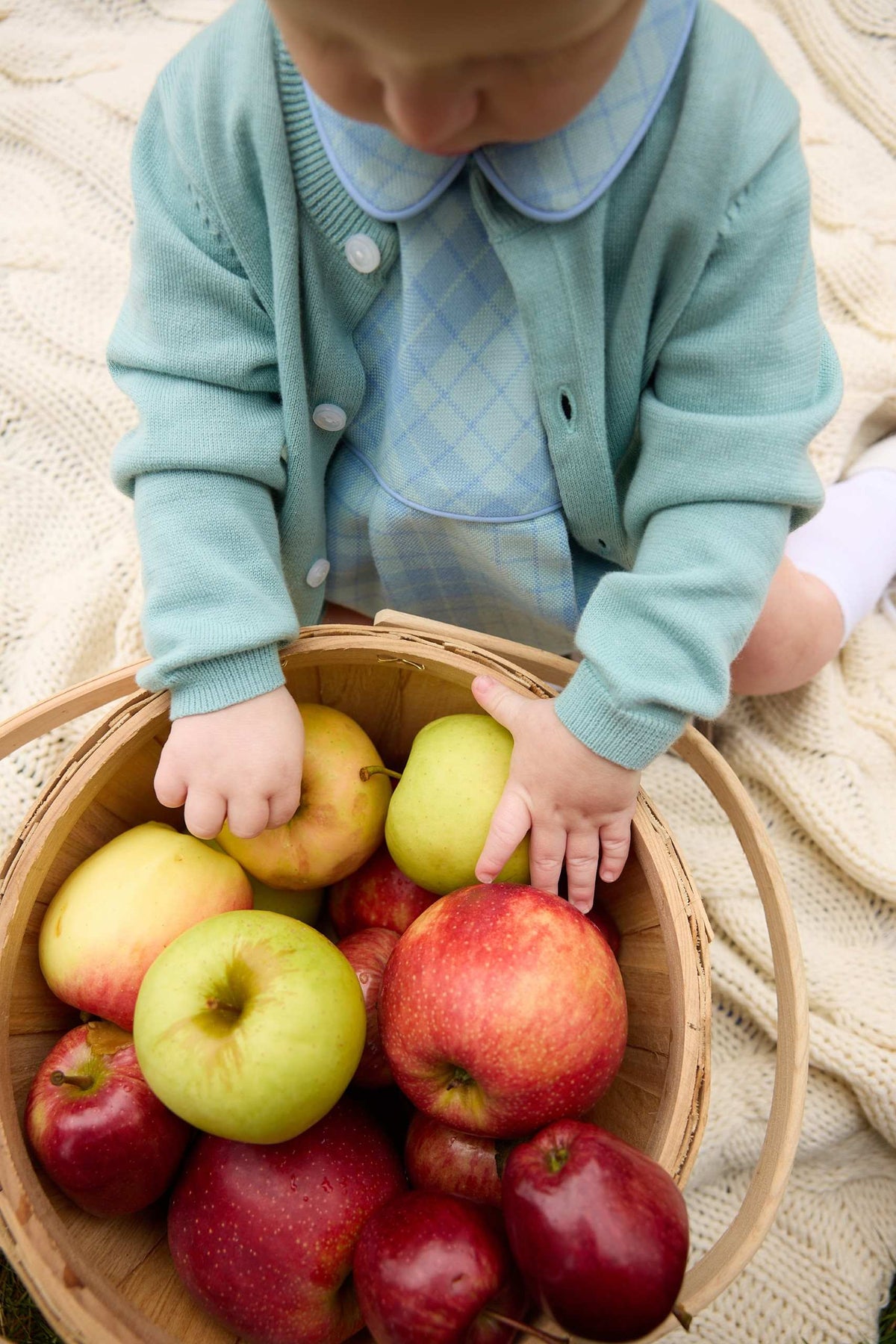 A young child in a blue outfit sits on a blanket, cozily wrapped in The Beaufort Bonnet Company’s Baby Girls’ and Girls’ Cambridge Cardigan with pearlized buttons, while holding a basket filled with red and green apples.