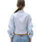 A person stands facing away, wearing the Simkhai Boone Combo Shirt—a light blue and white striped cropped shirt with rolled sleeves—paired with high-waisted blue jeans.