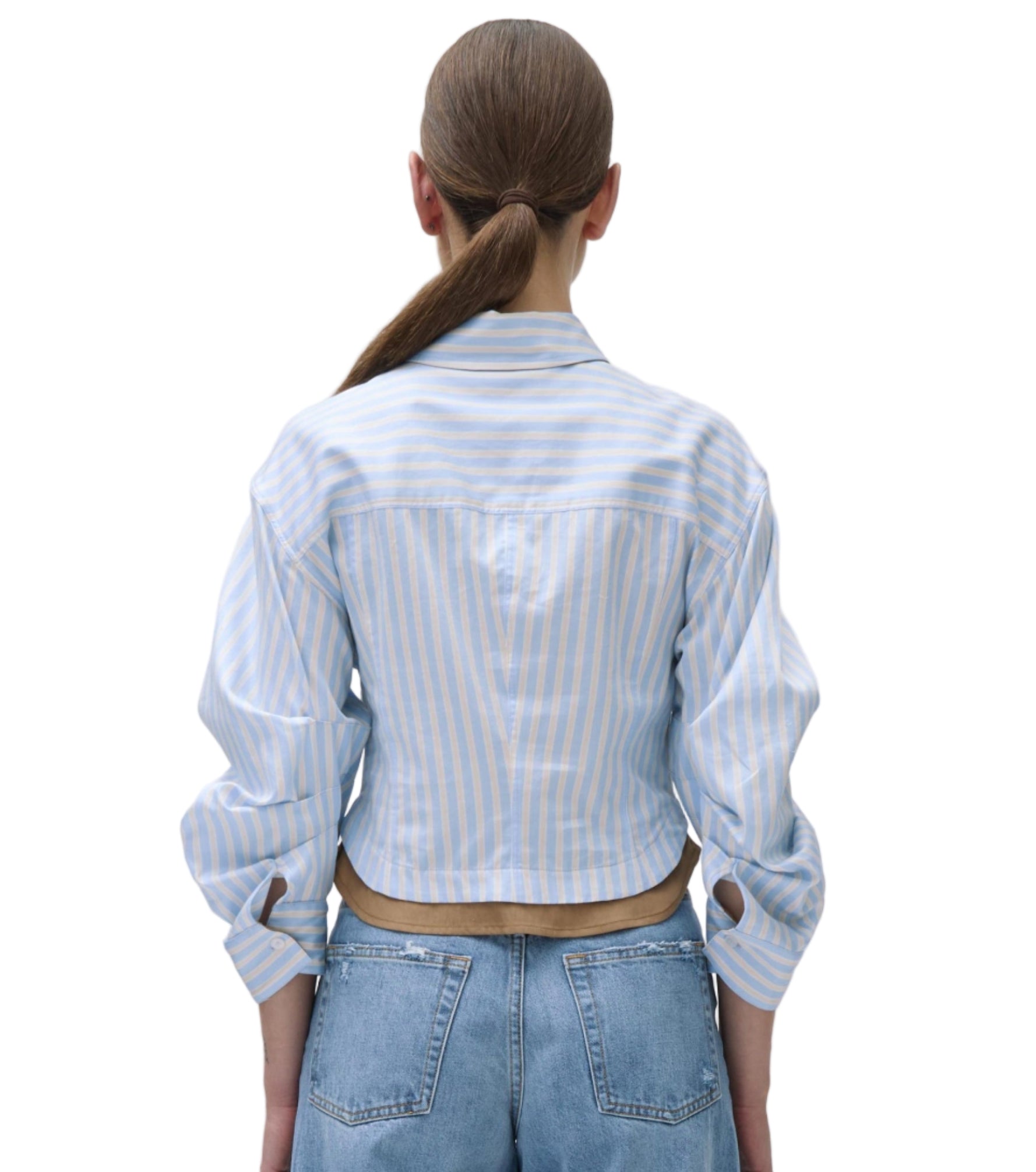 A person stands facing away, wearing the Simkhai Boone Combo Shirt—a light blue and white striped cropped shirt with rolled sleeves—paired with high-waisted blue jeans.