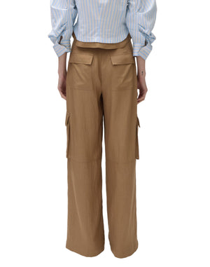 A person is seen from behind wearing Simkhai’s Ivrie Utility Pant—light brown, wide-leg cargo pants with back and side flap pockets—paired with a light blue striped long-sleeve shirt.