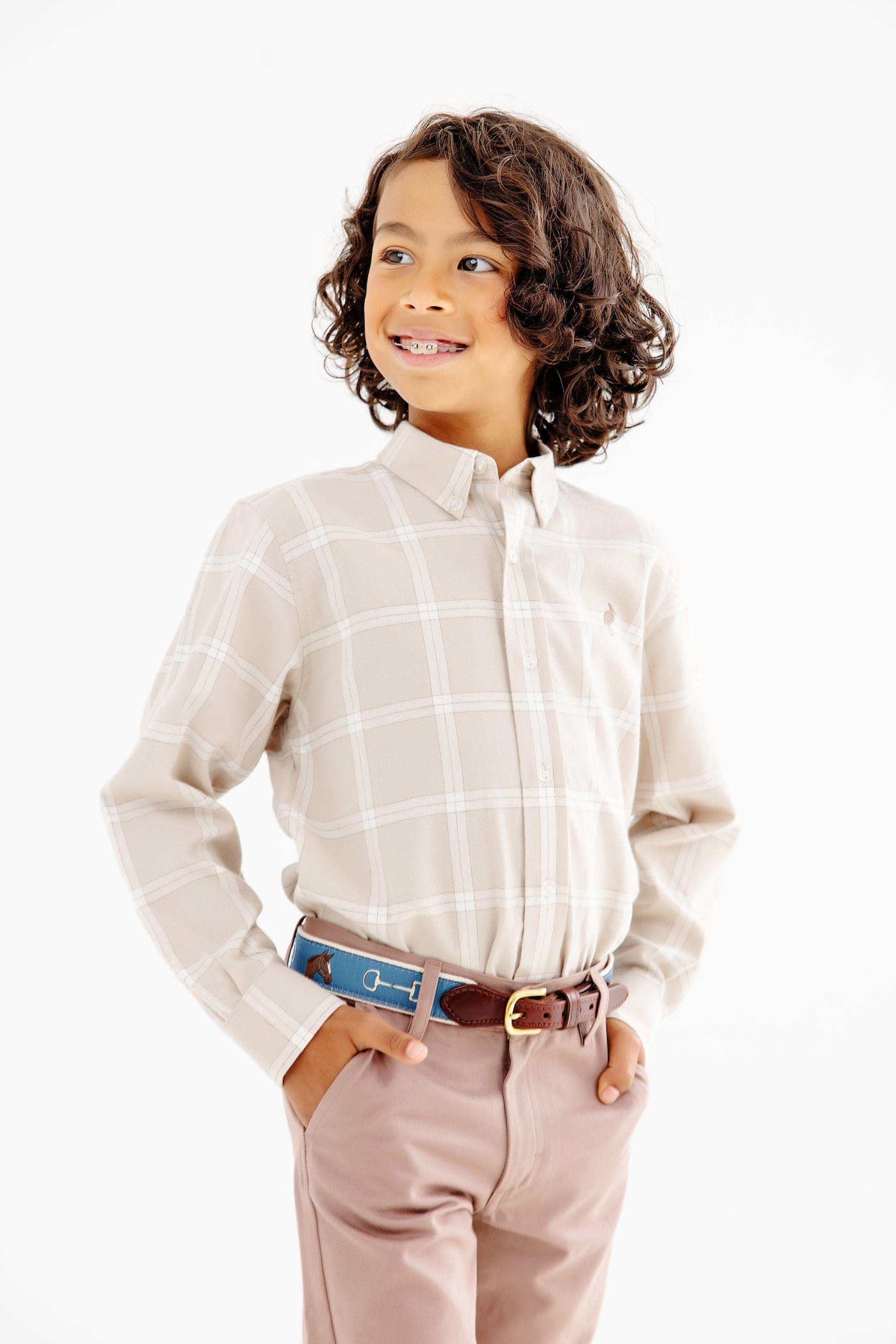 A young child with curly hair, wearing The Beaufort Bonnet Company Boys’ Flannel Dean’s List Dress Shirt, tan pants, and a belt, stands with hands in pockets, smiling and looking to the side against a white background.