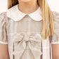 A young girl smiles while wearing the Baby Girls’ and Girls’ Francene Frock by The Beaufort Bonnet Company, a beige plaid dress featuring a white Peter Pan collar and a large bow on the chest.