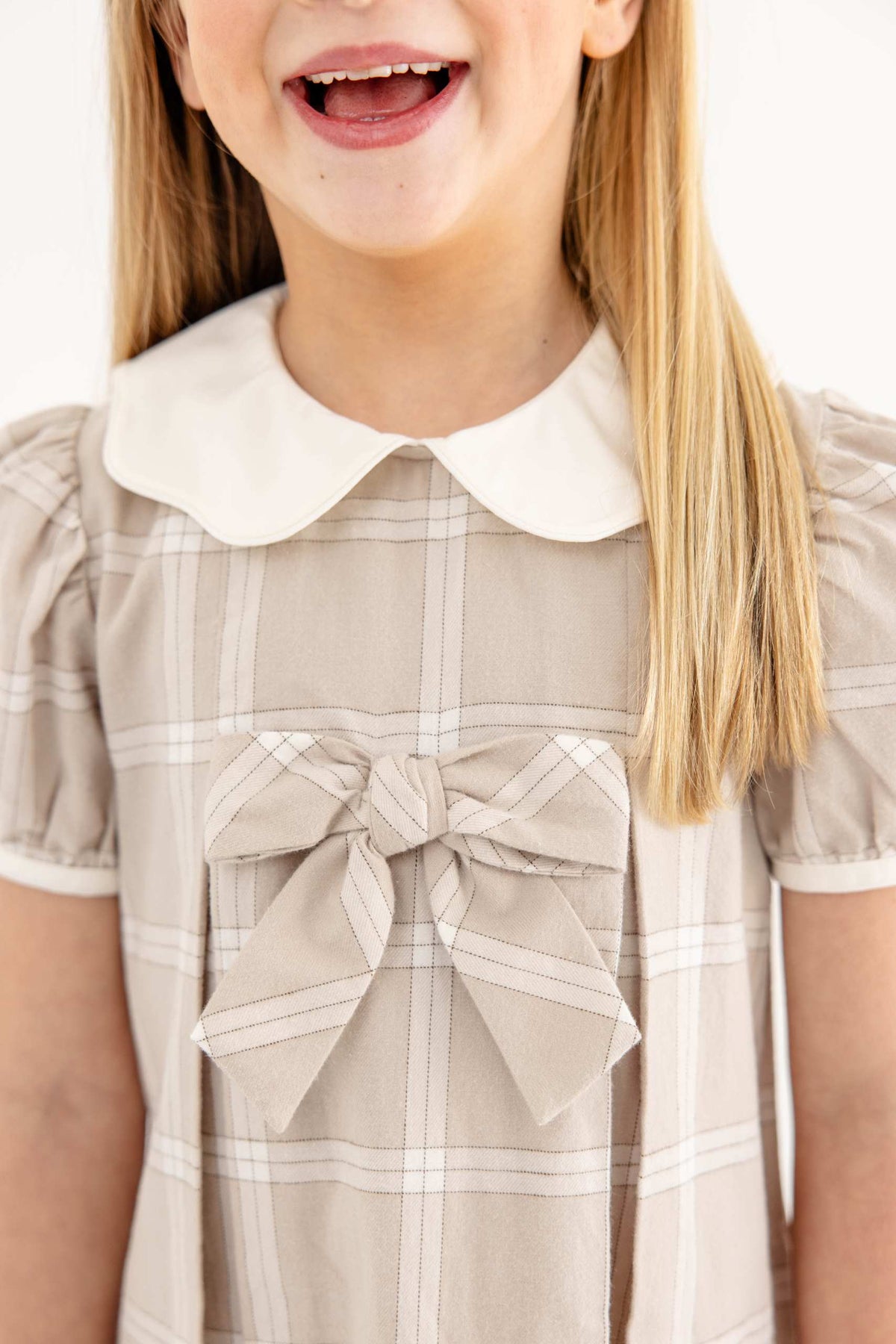 A young girl smiles while wearing the Baby Girls’ and Girls’ Francene Frock by The Beaufort Bonnet Company, a beige plaid dress featuring a white Peter Pan collar and a large bow on the chest.