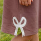 A child’s hand rests on The Beaufort Bonnet Company Girls’ Juliet Jumper in mauve, featuring a white bow applique near the hem; legs and grass can be seen.