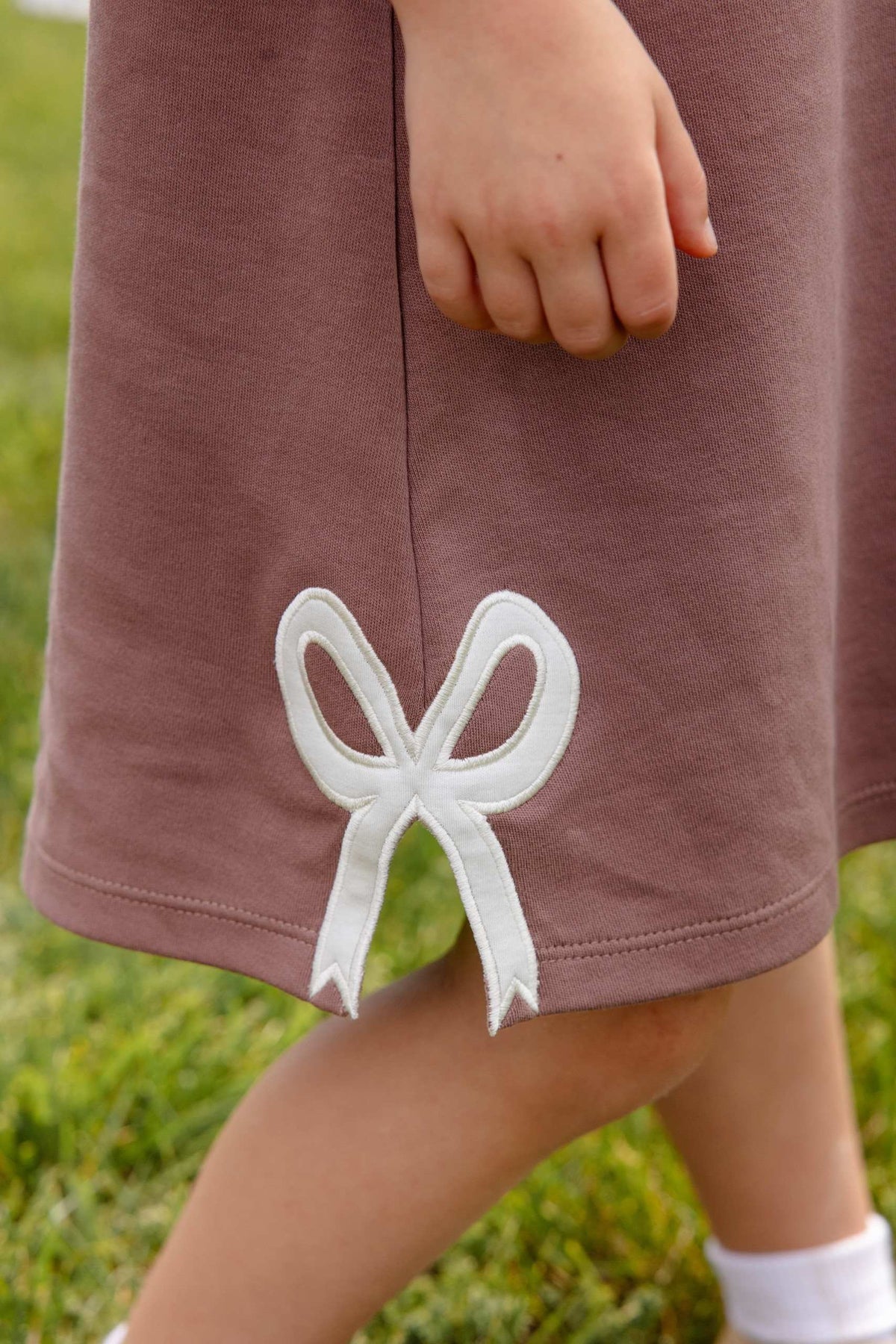 A child’s hand rests on The Beaufort Bonnet Company Girls’ Juliet Jumper in mauve, featuring a white bow applique near the hem; legs and grass can be seen.