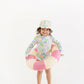 A young child, smiling at the camera, stands barefoot inside a pink and white swim ring against a plain white background, wearing The Beaufort Bonnet Company’s Baby Girls’ and Girls’ Sarasota Surf Suit along with a coordinating hat. This UPF 35+ swimsuit offers both stylish design and reliable sun protection for your little one.