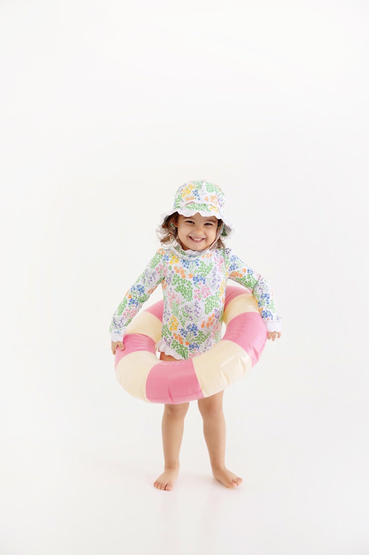 A young child, smiling at the camera, stands barefoot inside a pink and white swim ring against a plain white background, wearing The Beaufort Bonnet Company’s Baby Girls’ and Girls’ Sarasota Surf Suit along with a coordinating hat. This UPF 35+ swimsuit offers both stylish design and reliable sun protection for your little one.