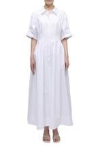 A person wearing the Simkhai Mollie Shirt Dress—a long, white, button-down dress with short sleeves and a collar—stands in white heeled sandals against a plain white background.