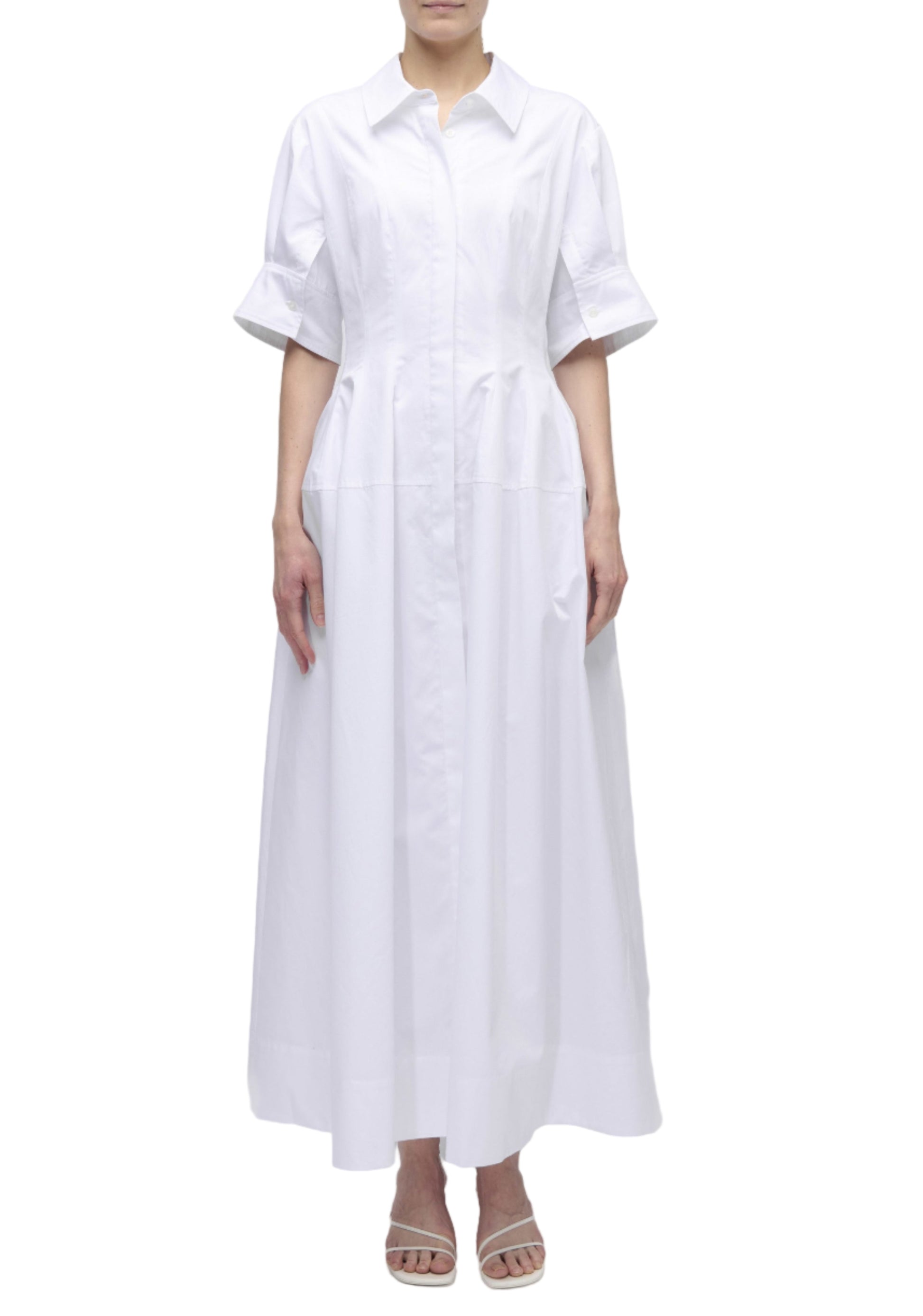 A person wearing the Simkhai Mollie Shirt Dress—a long, white, button-down dress with short sleeves and a collar—stands in white heeled sandals against a plain white background.