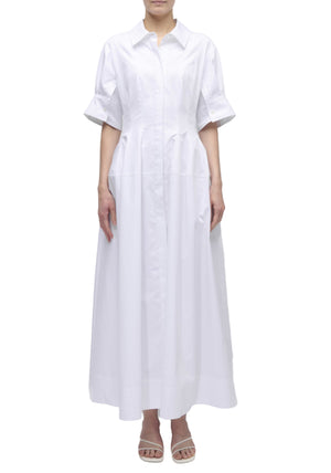 A person wearing the Simkhai Mollie Shirt Dress—a long, white, button-down dress with short sleeves and a collar—stands in white heeled sandals against a plain white background.